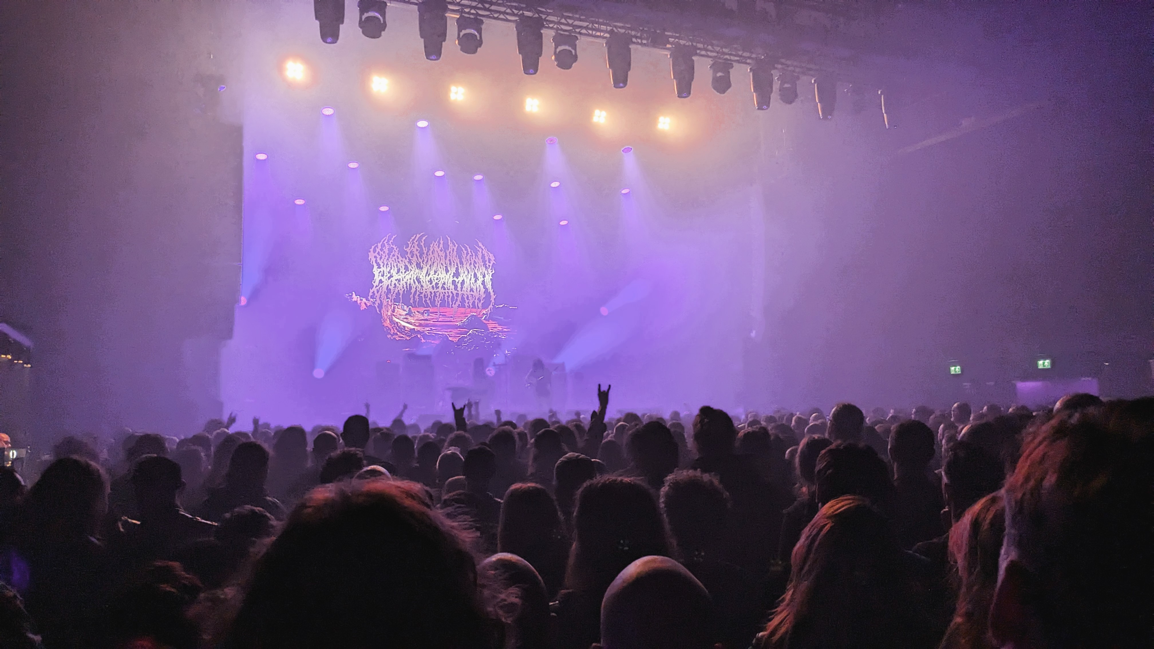 A crowded concert venue with a vibrant stage, illuminated by purple and yellow lights, as attendees enjoy a performance.