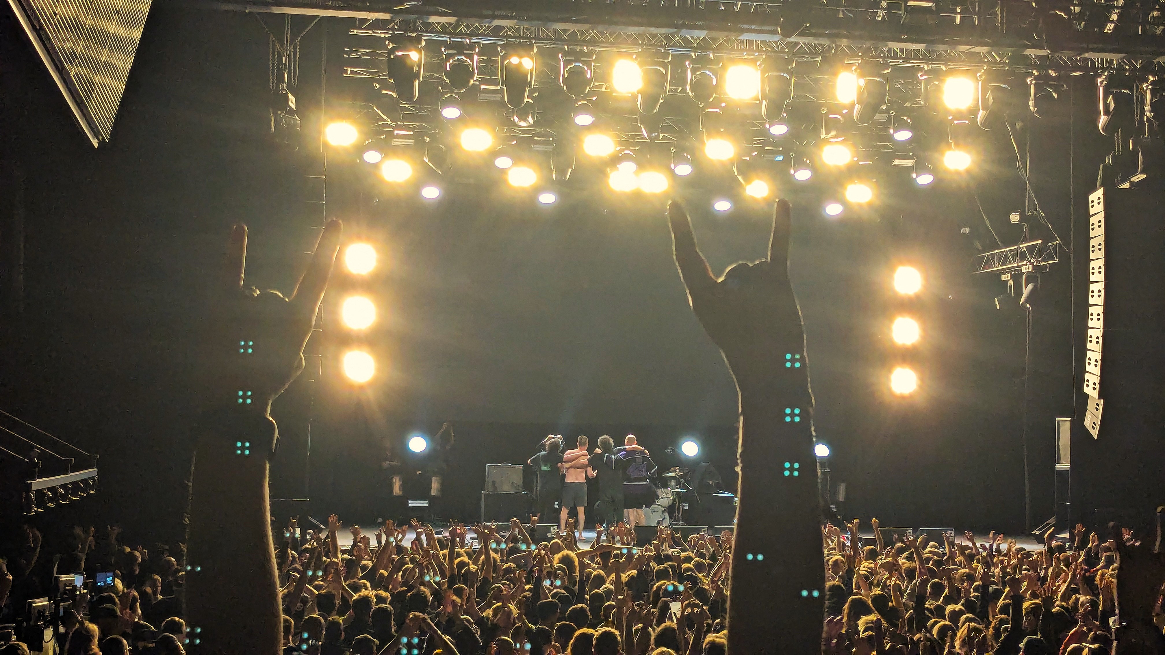 A large crowd of concert-goers raises their hands in the air, making the rock 'n' roll gesture with their fingers, in front of a brightly lit stage during a music festival.