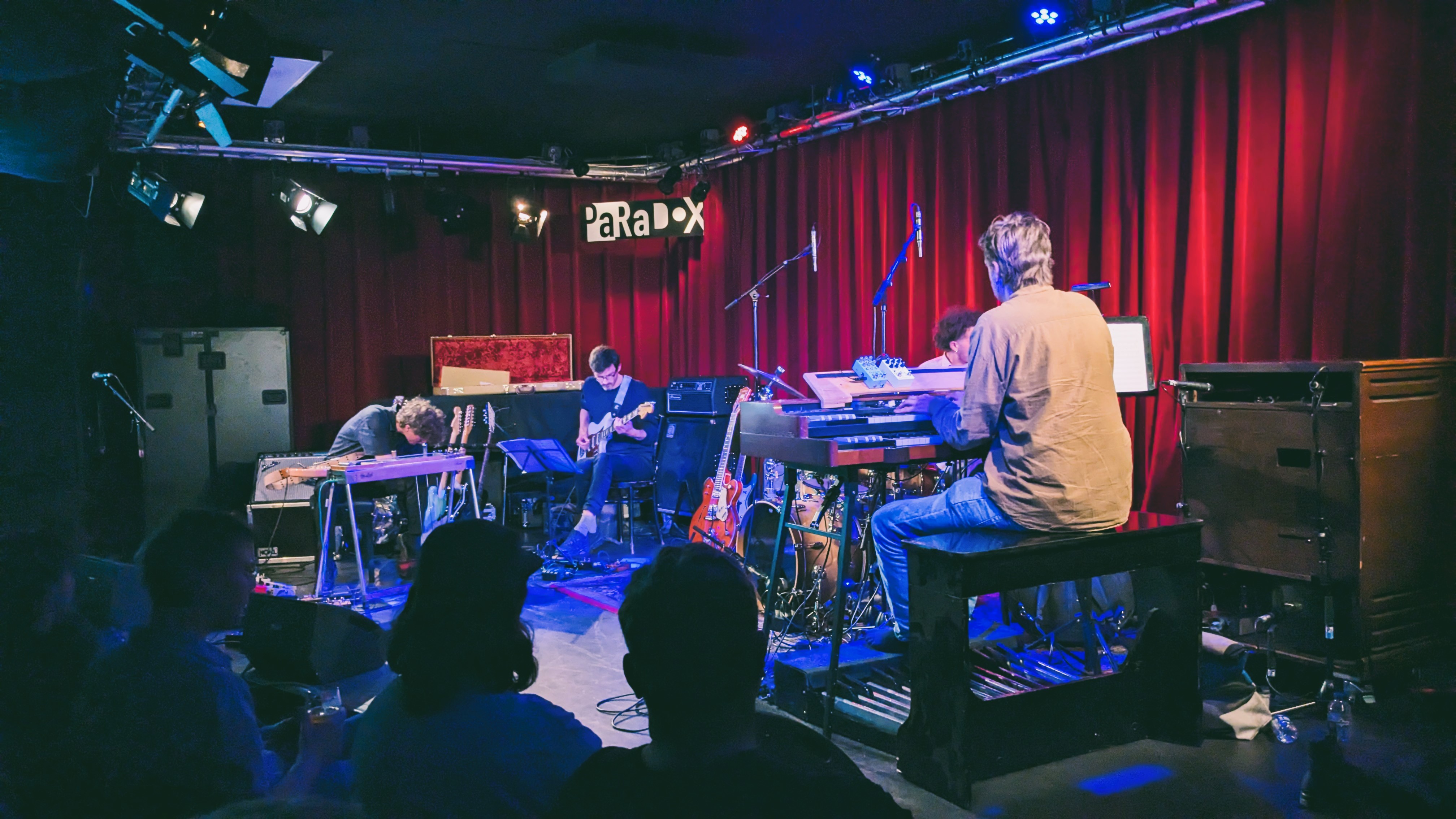 A live music performance in a small venue featuring musicians playing various instruments on stage, with red curtains in the background.