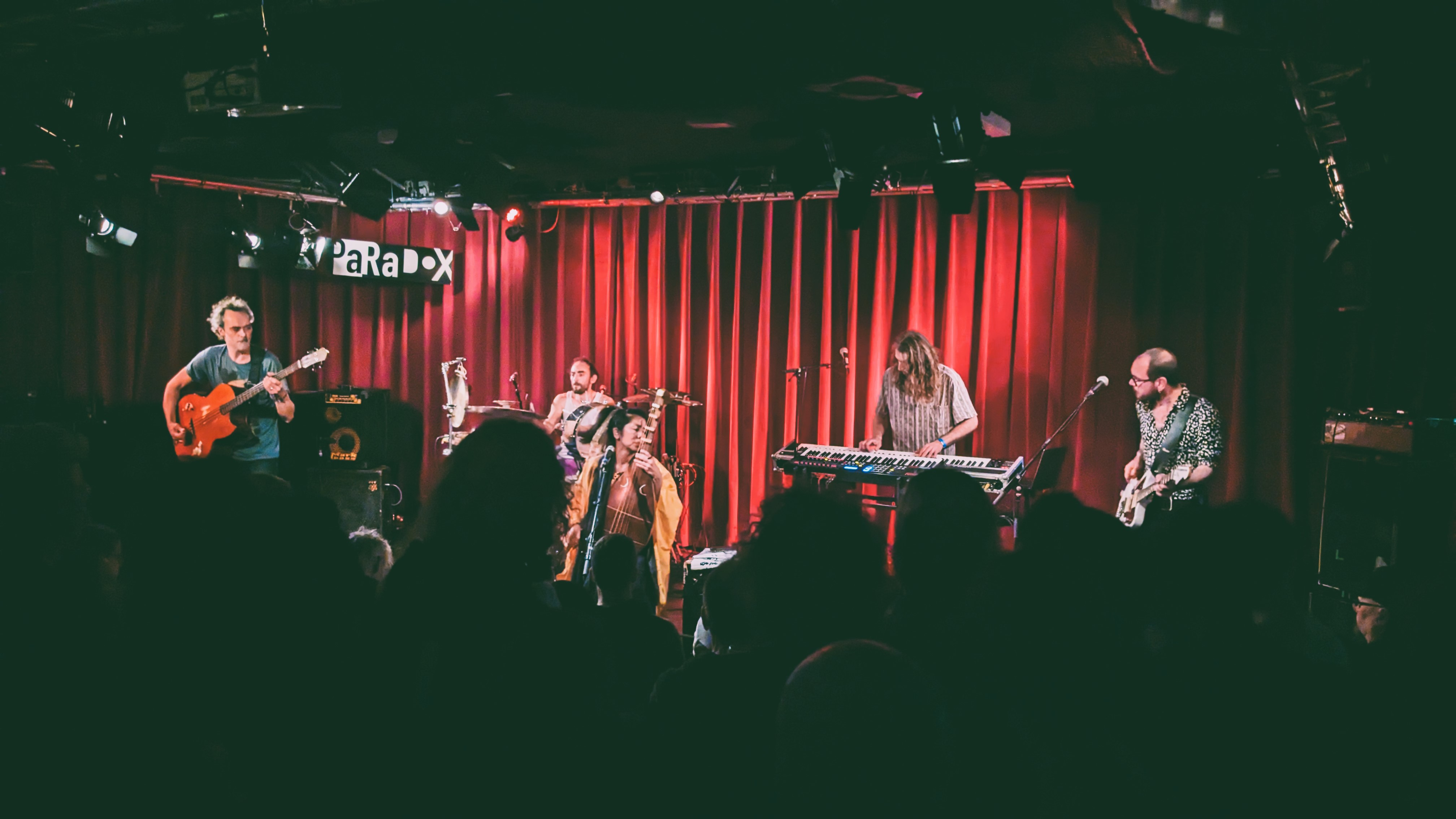 A live music performance featuring a diverse band on stage, with a guitarist, keyboardist, and another musician, in a warmly lit venue decorated with red curtains.