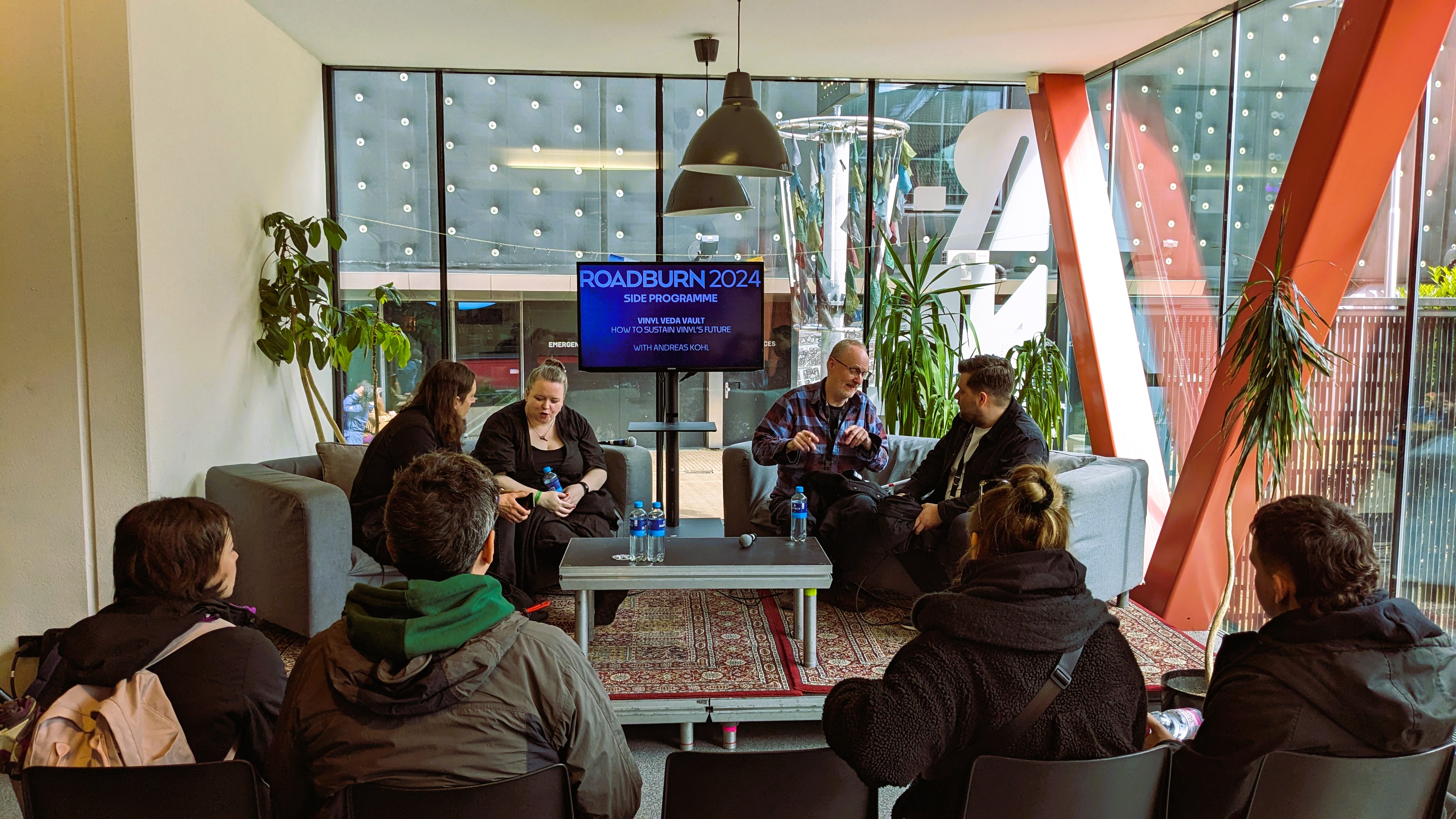 A panel discussion at the Roadburn 2024 festival featuring speakers seated at a table, with an audience in view, and a large screen displaying the event details in the background.