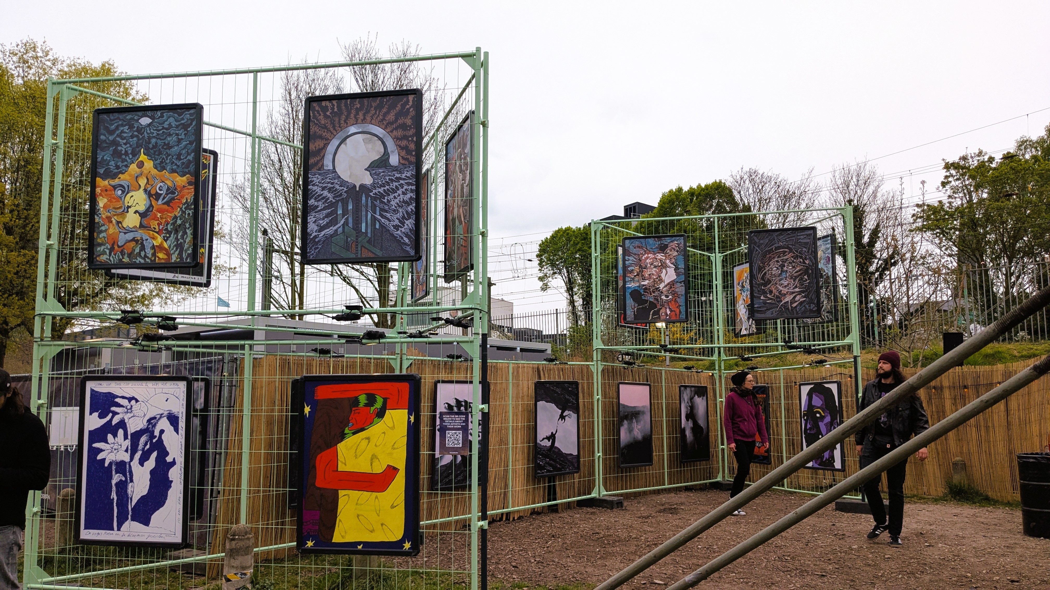 An outdoor exhibition featuring colorful framed artworks displayed on a metal structure, with attendees observing the pieces.