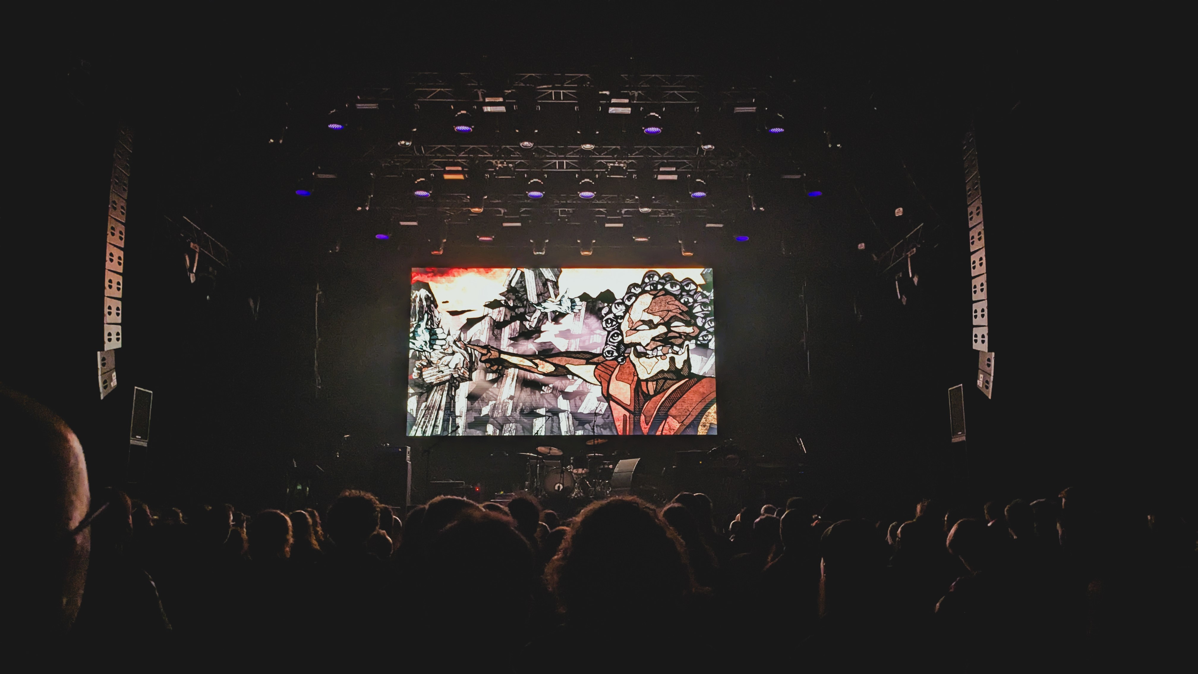 A crowded concert venue with an audience watching a large screen displaying animated artwork, featuring a character with a dramatic expression and pointing gesture.