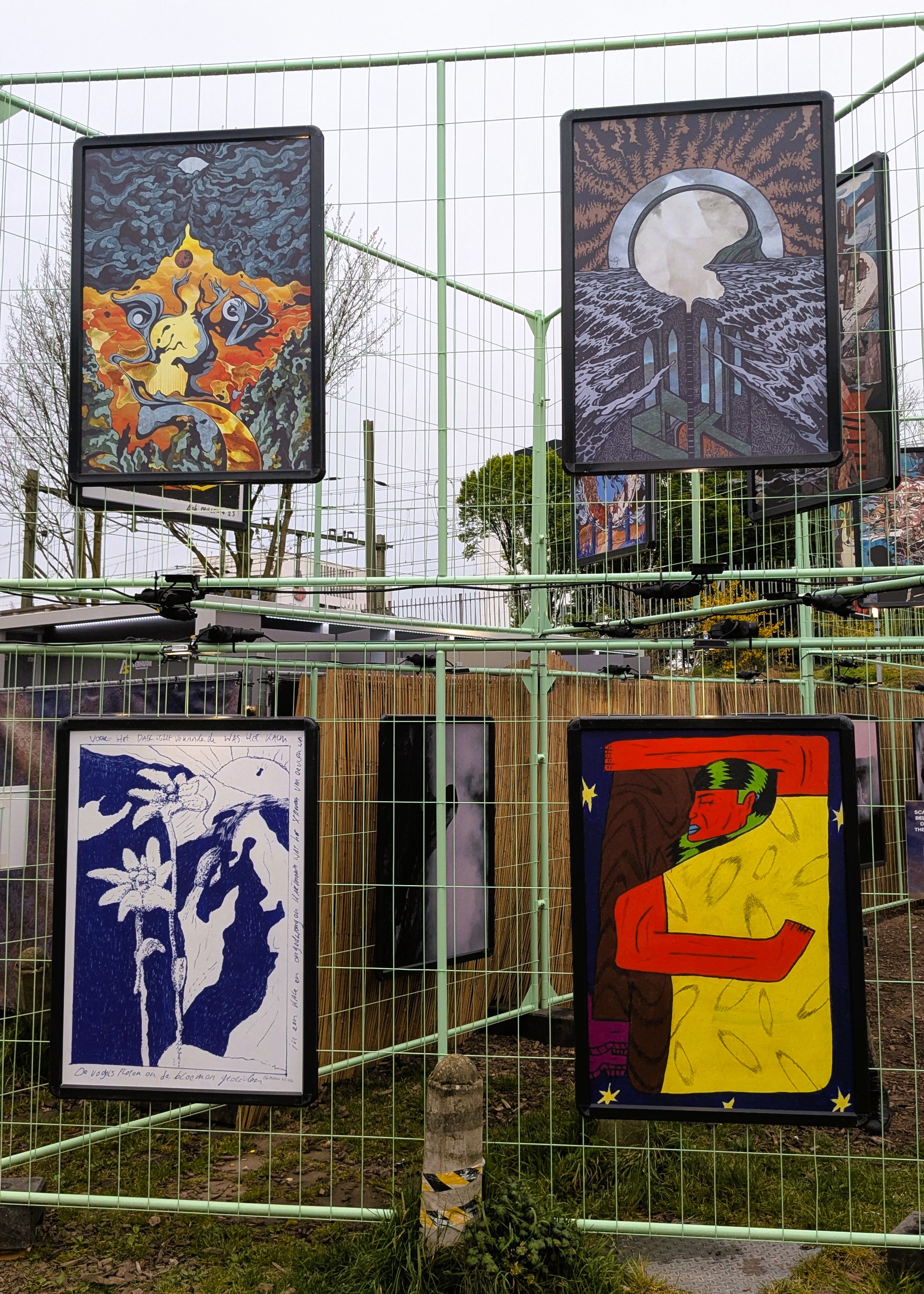 An outdoor exhibition featuring colorful artwork displayed on panels inside a green fence, with various artistic styles and themes represented.