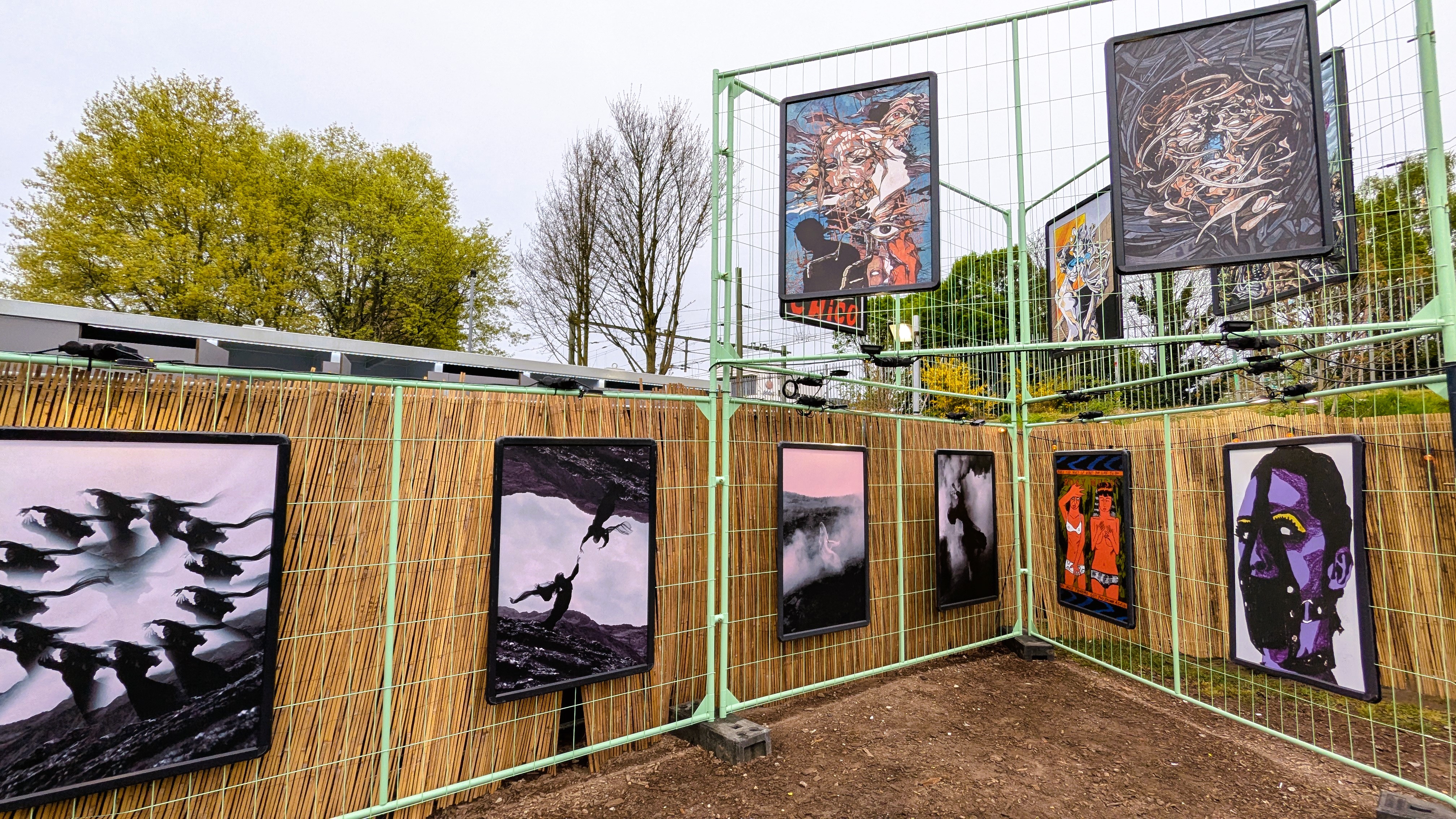 A display of artistic posters at an outdoor exhibition, featuring various styles and themes, surrounded by a green fence and trees in the background.
